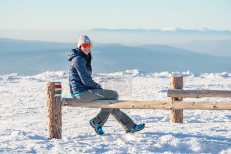 Photo for Young woman sits on a wooden stump on top of a mountain and rests after hiking on a winter mountain trail, mountain landscape in winter. - Royalty Free Image
