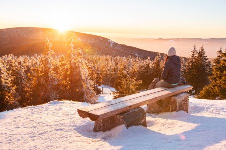 Sudetes, una mujer joven se sienta en un banco con vistas al paisaje de montaña al atardecer, vista desde una ruta de senderismo de invierno.