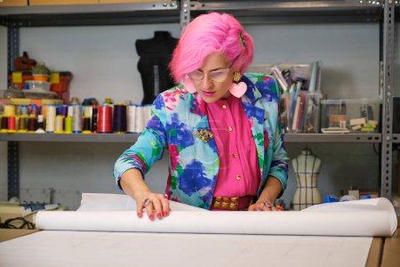 Photo for Seamstress with pink hair and colorfull clothes unrolling sewing patterns in a sewing workshop. - Royalty Free Image