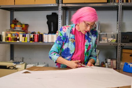 Photo for Tailor with pink hair and colorfull clothes cutting sewing patterns in a sewing workshop. - Royalty Free Image