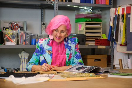Photo for Dressmaker with pink hair and colorfull clothes taking ideas from fashion magazines in a sewing workshop. - Royalty Free Image