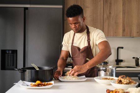 Téléchargez les photos : Jeune homme noir servant du poulet frit haché sur du riz dans une assiette dans une cuisine. - en image libre de droit