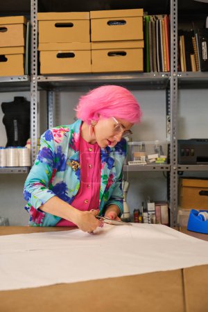 Photo for Seamstress with pink hair and colorfull clothes cutting sewing patterns in a sewing workshop. - Royalty Free Image