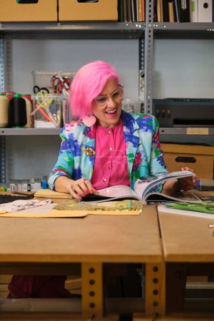 Photo for Tailor with pink hair and colorfull clothes taking ideas from fashion magazines in a sewing workshop. - Royalty Free Image