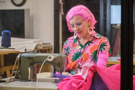 Photo for Dressmaker with pink hair and colorfull clothes smiling and looking at camera working on an industrial sewing machine in a sewing workshop. - Royalty Free Image