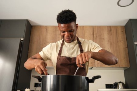 Téléchargez les photos : Young african man cooking in a big pot in a kitchen. - en image libre de droit