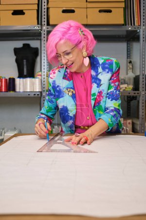 Photo for Dressmaker with pink hair and colorfull clothes drawing sewing patterns in a sewing workshop. - Royalty Free Image