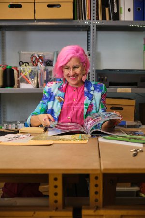 Photo for Seamstress with pink hair and colorfull clothes taking ideas from fashion magazines in a sewing workshop. - Royalty Free Image
