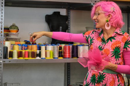 Photo for Tailor with pink hair and colorfull clothes choosing a thread in a sewing workshop. - Royalty Free Image