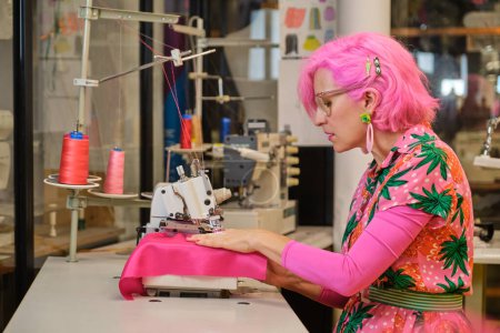Photo for Dressmaker with pink hair and colorfull clothes working on an overlocker in a sewing workshop. - Royalty Free Image