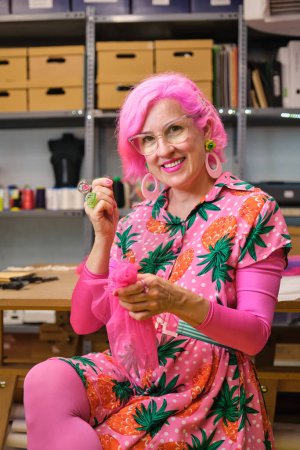 Photo for Dressmaker with pink hair and colorfull clothes hand sewing a piece of fabric, smiling and looking at camera, in a sewing workshop. - Royalty Free Image