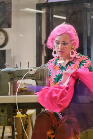Photo for Seamstress with pink hair and colorfull clothes smiling and looking at camera working on an industrial sewing machine in a sewing workshop. - Royalty Free Image