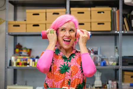 Photo for Seamstress with pink hair and colorfull clothes making funny faces with sewing thread reels in a sewing workshop. - Royalty Free Image