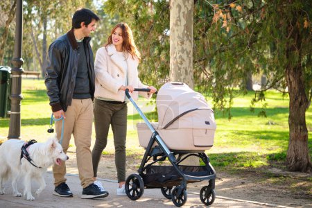 Man and pregnant woman walking with their dog and baby stroller in a park. Family.