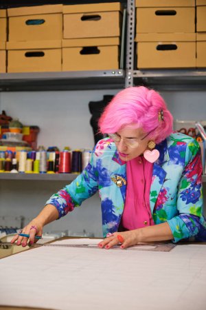 Photo for Seamstress with pink hair and colorfull clothes drawing sewing patterns in a sewing workshop. - Royalty Free Image