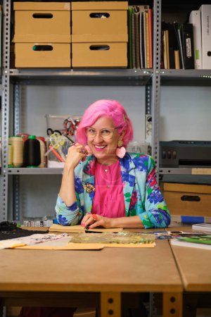 Photo for Seamstress with pink hair and colorfull clothes smiling and looking at camera in a sewing workshop. - Royalty Free Image
