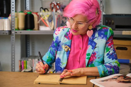 Photo for Seamstress with pink hair and colorfull clothes drawing new layout in a sewing workshop. - Royalty Free Image
