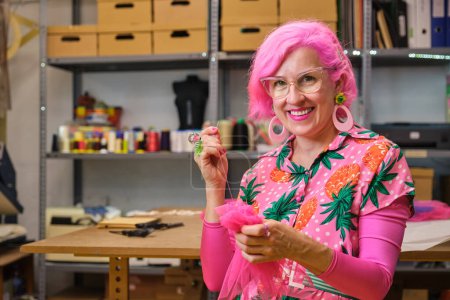 Photo for Seamstress with pink hair and colorfull clothes hand sewing a piece of fabric, smiling and looking at camera, in a sewing workshop. - Royalty Free Image