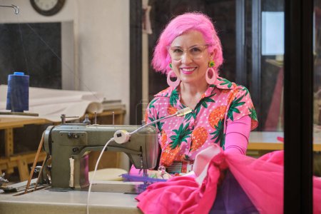 Photo for Tailor with pink hair and colorfull clothes smiling and looking at camera working on an industrial sewing machine in a sewing workshop. - Royalty Free Image