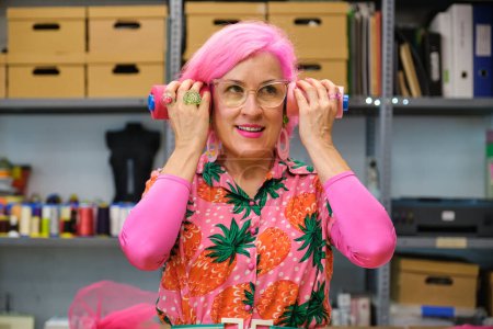 Photo for Tailor with pink hair and colorfull clothes making funny faces with sewing thread reels in a sewing workshop. - Royalty Free Image