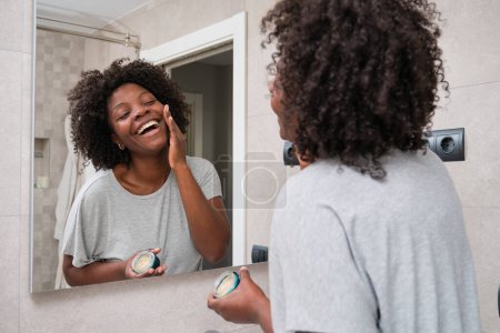 Smiling african young woman applying eco-friendly facial cream in bathroom.