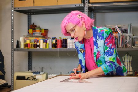 Photo for Tailor with pink hair and colorfull clothes drawing sewing patterns in a sewing workshop. - Royalty Free Image