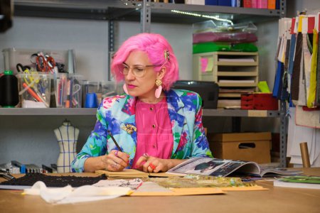 Photo for Tailor with pink hair and colorfull clothes taking ideas from fashion magazines in a sewing workshop. - Royalty Free Image