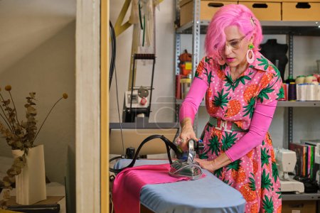 Photo for Tailor with pink hair and colorfull clothes ironing fabric in a sewing workshop. - Royalty Free Image