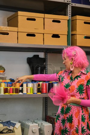 Photo for Dressmaker with pink hair and colorfull clothes choosing a thread in a sewing workshop. - Royalty Free Image