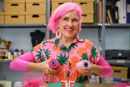 Photo for Dressmaker with pink hair and colorfull clothes making funny faces with sewing thread reels in a sewing workshop. - Royalty Free Image