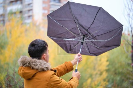 Jeune homme essaie de tenir son parapluie dans un vent fort par un jour d'automne pluvieux dans la rue.