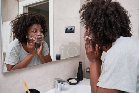 Téléchargez les photos : Femme africaine maintient sa beauté avec la routine quotidienne de lavage du visage en utilisant du savon dans la salle de bain. - en image libre de droit