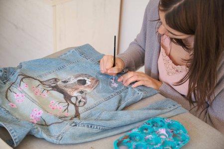 Caucasian woman hand painting a design using acrylic paints of a deer and flowers in a denim jacket. Unique clothing designs.