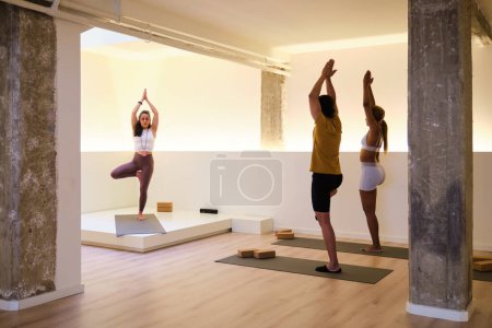 A group of people are practicing Vrksasana or tree yoga pose in a room. Yoga class.