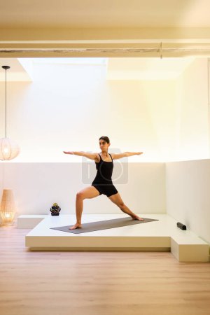 Eine Frau macht in einem Raum mit weißer Wand und Holzboden Virabhadrasana II oder Krieger II Yoga-Pose. Der Raum ist schwach beleuchtet, und es gibt eine Lampe auf dem Boden.