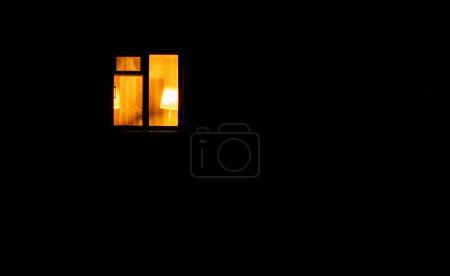Foto de La ventana solitaria en una habitación brilla en la noche oscura. La luz amarilla brillante arde sobre el fondo negro. Vida nocturna solitaria. Concepto abstracto uno entre el resto. Apagón. - Imagen libre de derechos