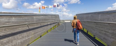 Téléchargez les photos : Delfzijl, Pays-Bas - 8 août 2023 : Touriste féminine avec sac à dos traversant le pont vers la plate-forme d'observation et promenade le long de la plage de Delfzijl dans la province de Groningue aux Pays-Bas - en image libre de droit