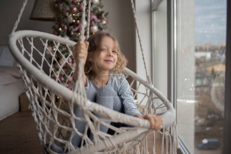 Photo for Child happiness with present in room and x-mas tree and sits on the swing against the window. High quality photo - Royalty Free Image