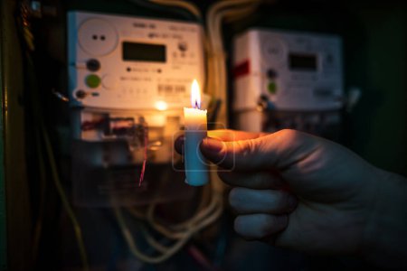 Kerzen leuchten im Dunkeln in der Nähe von Stromzählern während eines Stromausfalls zu Hause. kein Strom. Hand hält eine brennende Kerze.