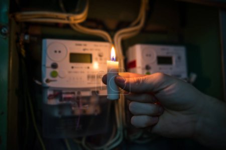 Kerzen leuchten im Dunkeln in der Nähe von Stromzählern während eines Stromausfalls zu Hause. kein Strom. Hand hält eine brennende Kerze.