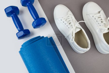 Foto de Primer plano de una estera de yoga de color azul enrollada, zapatillas blancas, pesas, estilo de vida saludable, deporte y concepto de ejercicio. Fondo gris. - Imagen libre de derechos
