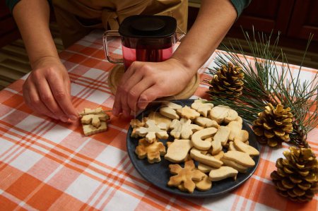Foto de Enfoque selectivo en un ama de casa usando cuerda de lino, atando galletas de jengibre. Rama de abeto, conos de pino dorado como adorno de Navidad en una mesa de cocina con un plato lleno de pasteles festivos caseros - Imagen libre de derechos