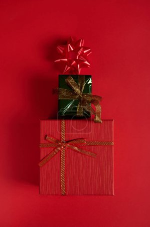 Foto de Acostado. Vertical studio shot de lindas cajas de regalo elegantes, dispuestas en forma de árbol de Navidad, decoradas con lazo sobre fondo rojo. Naturaleza muerta. Evento de Año Nuevo. Día de Boxeo. 25 de diciembre. Navidad. - Imagen libre de derechos