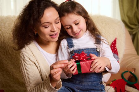 Photo for Close up portrait of beautiful loving mother - multi-ethnic woman, tenderly hugging her adorable little daughter unpacking Christmas present. Winter New Year holidays. Happy childhood and motherhood - Royalty Free Image