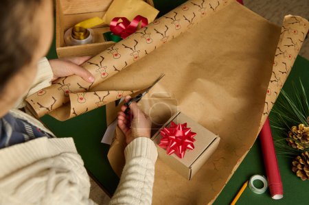 Photo for Overhead view of an unrecognizable woman, using scissors, cuts the decorative wrapping paper for packing gift for Christmas, New Year or other celebration event. Handsworkart. Boxing Day. Diy presents - Royalty Free Image