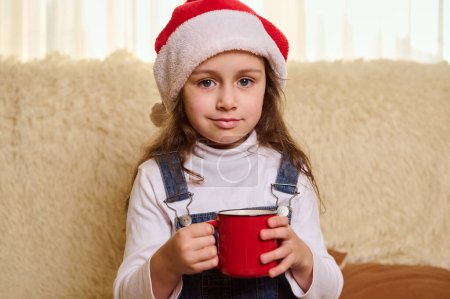 Foto de Retrato de una niña en el sombrero de Santa, sonriendo mirando a la cámara, disfrutando de una deliciosa bebida dulce de chocolate caliente con malvaviscos, en el acogedor interior de la casa. Felices fiestas de invierno. Temas de Navidad - Imagen libre de derechos