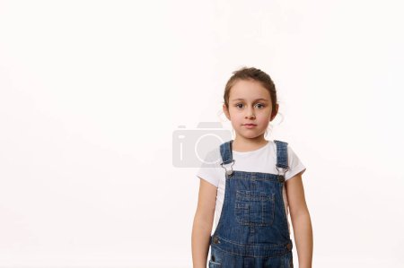 Foto de Retrato seguro de una adorable niña caucásica, vistiendo una camiseta blanca y un mono de mezclilla azul, mirando a la cámara, aislada sobre fondo blanco con espacio para copiar texto promocional. - Imagen libre de derechos