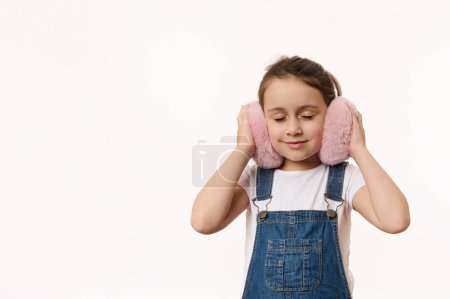 Téléchargez les photos : Adorable enfant caucasien, jolie petite fille à la mode portant des salopettes en denim, se réchauffant les oreilles avec son nouvel accessoire de vêtements moelleux rose à la mode - des cache-oreilles moelleux. Copier l'espace sur fond blanc - en image libre de droit