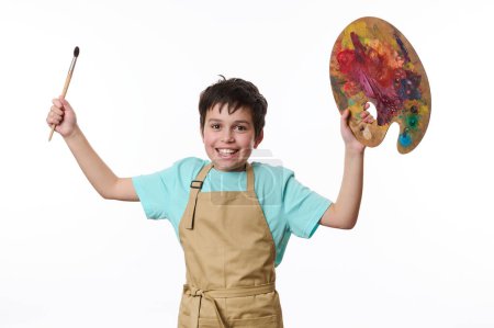Photo for Mischievous teenage boy wearing beige apron, talented painter with paintbrush and palette of colorful paints, cutely smiling a toothy smile looking at camera, white background. Kids art development - Royalty Free Image