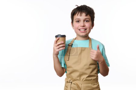 Téléchargez les photos : Portrait isolé sur fond blanc d'un jeune garçon barman heureux montrant pouce levé, vendant une boisson à café à emporter dans une tasse en carton. Boisson chaude à emporter. Espace de copie pour votre publicité - en image libre de droit